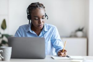 A woman sat at a desk with a headset on her head in front of a laptop, whilst writing with a pen.
